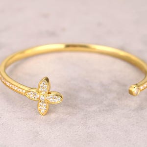 Op de afbeelding: Een open manchetarmband in goudkleur. De armband heeft een vierbladige bloemvorm met heldere, sprankelende edelstenen in het midden en op de bloemblaadjes. De armband heeft een gladde, gepolijste afwerking en staat tegen een lichtgrijze achtergrond.