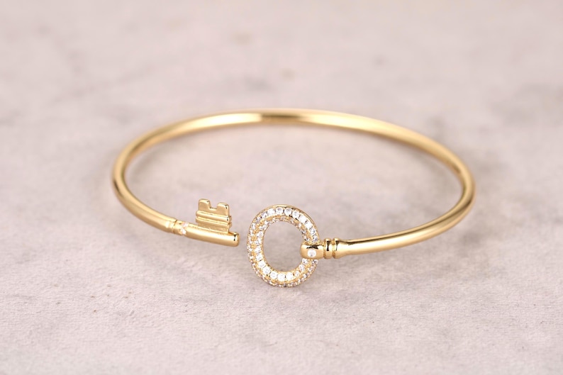 Puede incluir: Una pulsera dorada con un dise&ntilde;o de llave. La cabeza de la llave es un c&iacute;rculo incrustado con peque&ntilde;as piedras preciosas transparentes. La pulsera est&aacute; hecha de un metal liso y brillante y est&aacute; abierta en un extremo, con la cabeza de la llave y los dientes de la llave formando el cierre.