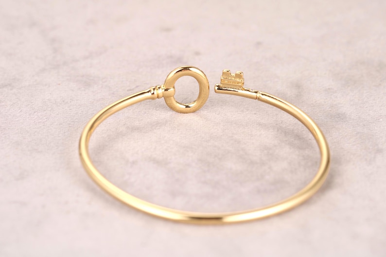 Puede incluir: Un brazalete dorado en forma de llave. El brazalete presenta un dise&ntilde;o de llave en el cierre, con la cabeza y el asa circular de la llave formando el cierre. El brazalete tiene aproximadamente 6,35 cm de di&aacute;metro.