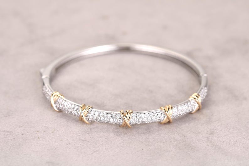May include: A silver-tone bracelet with a central band encrusted with small, clear gemstones. The band is accented with gold-tone X-shaped designs. The bracelet is a hinged style, designed to be worn on the wrist. The jewelry is set against a neutral background.