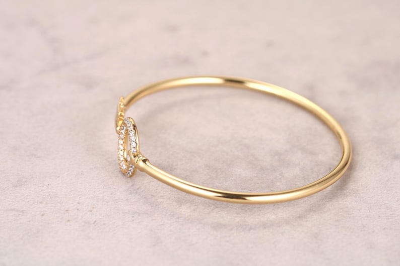 Puede incluir: Un brazalete dorado con un cierre decorativo. El cierre est&aacute; adornado con peque&ntilde;as piedras preciosas transparentes. El brazalete se encuentra sobre un fondo neutro, resaltando su elegante dise&ntilde;o. Una joya.