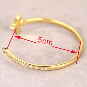 Puede incluir: Una pulsera dorada con un dise&ntilde;o floral y una fila de peque&ntilde;as piedras transparentes. La pulsera tiene 5 cm de di&aacute;metro. La pulsera est&aacute; sobre una superficie gris claro.