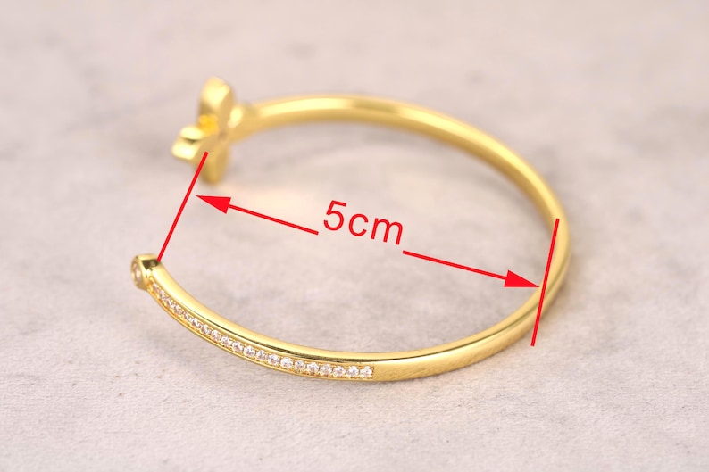 Puede incluir: Una pulsera dorada con un dise&ntilde;o floral y una fila de peque&ntilde;as piedras transparentes. La pulsera tiene 5 cm de di&aacute;metro. La pulsera est&aacute; sobre una superficie gris claro.