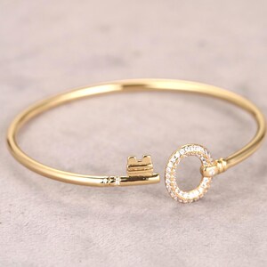 Puede incluir: Un brazalete dorado en forma de llave. La cabeza de la llave es un c&iacute;rculo incrustado con peque&ntilde;as piedras preciosas transparentes. El eje de la llave tiene un dise&ntilde;o simple y elegante. El brazalete est&aacute; sobre un fondo neutro.