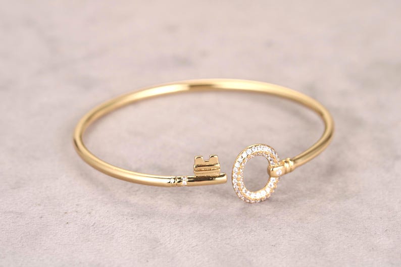 Puede incluir: Un brazalete dorado en forma de llave. La cabeza de la llave es un c&iacute;rculo incrustado con peque&ntilde;as piedras preciosas transparentes. El eje de la llave tiene un dise&ntilde;o simple y elegante. El brazalete est&aacute; sobre un fondo neutro.