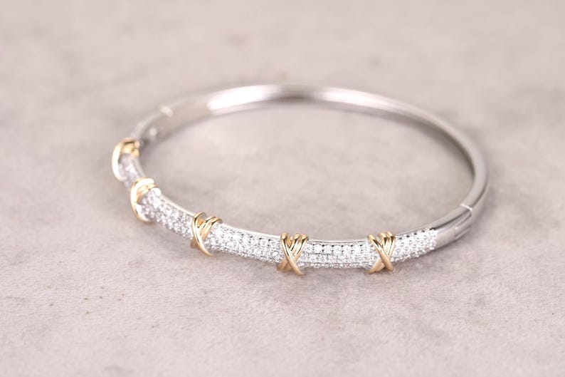 May include: Silver-toned bangle bracelet with a row of small, sparkling stones. The bracelet features gold-colored accents in an X-shaped design. The jewelry is displayed on a neutral gray surface.
