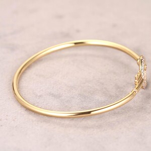 Puede incluir: Una pulsera de brazalete dorada con un acento decorativo enjoyado. La pulsera es circular y parece estar hecha de un material met&aacute;lico brillante. El acento presenta peque&ntilde;as piedras preciosas claras. El fondo es de color gris claro neutro.