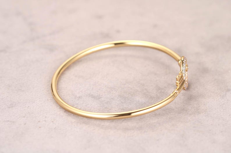 Puede incluir: Una pulsera de brazalete dorada con un acento decorativo enjoyado. La pulsera es circular y parece estar hecha de un material met&aacute;lico brillante. El acento presenta peque&ntilde;as piedras preciosas claras. El fondo es de color gris claro neutro.