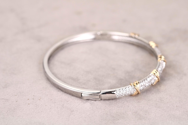 May include: A silver and gold-tone hinged bangle bracelet. The bracelet features a section of small, clear gemstones set in a silver band, accented with gold-tone details. The clasp is silver.
