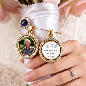 May include: A bridal bouquet of pink roses, with two gold memorial charms attached. One charm features a photo, the other has angel wings and the text "walking with you today and always."