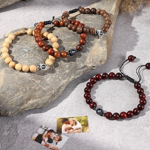 Puede incluir: Varios brazaletes de cuentas de madera en varios tonos de marrón y rojo se exhiben sobre una superficie de piedra. Los brazaletes presentan diferentes tamaños de cuentas y estilos de ensartado, algunos incorporando detalles metálicos. También hay dos fotos pequeñas de una pareja.