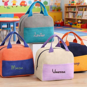 Puede incluir: Cuatro loncheras de pana en varios colores: naranja, gris, crema y azul marino. Cada bolsa tiene un panel de color contrastante con un nombre bordado. Las bolsas tienen asas y cremalleras de colores a juego. El fondo muestra un aula.