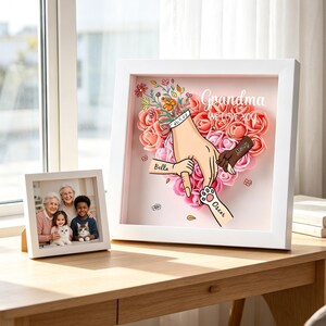 Puede incluir: Una caja de sombra con marco blanco con un diseño de corazón floral y las palabras "Grandma we love you". La obra de arte incluye manos, una huella de pata y los nombres "Bella" y "Oscar". Una foto enmarcada de una familia está al lado.