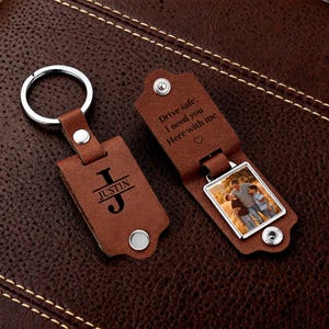 Puede incluir: Un llavero de cuero marrón con un llavero plateado. El llavero tiene el nombre "JUSTIN" y la inicial "J" grabada. El otro lado tiene una foto y el texto "Drive safe. I need you Here with me".