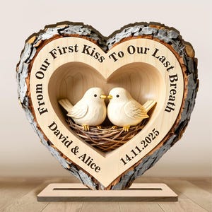 Puede incluir: Un adorno decorativo de madera en forma de corazón con borde de corteza. En el interior, dos pájaros blancos tallados se miran en un nido. El texto dice "From Our First Kiss To Our Last Breath" e incluye nombres y una fecha.