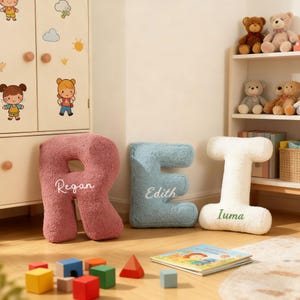 May include: A nursery scene featuring large, plush letter pillows spelling out "REI". The letters are in pink, blue, and white, with names embroidered on them. A children's book and wooden blocks are scattered on the floor, with stuffed animals on a shelf.