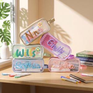 Puede incluir: Cinco estuches rectangulares transparentes con cremalleras y asas. Cada estuche presenta un nombre bordado en hilo en la parte frontal, incluyendo Nina, Wills, Emily, Sophia y Lycha, algunos decorados con pequeñas flores de hilo tejidas.