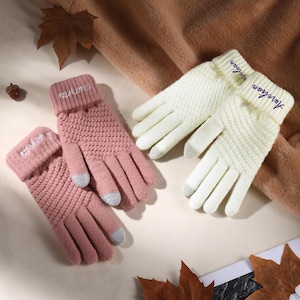 Peut inclure: Deux paires de gants tricotés en rose et crème. Les gants ont des bouts de doigts gris pour écran tactile et le mot "Aurelian" ou "Marguerite" brodé sur les poignets. Les gants sont présentés sur une surface neutre avec des feuilles d'automne.