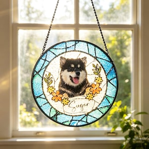 Puede incluir: Vidriera redonda que representa un perro negro y marrón con la lengua fuera, rodeado por una corona floral. El nombre del perro, "Sugar", está escrito en cursiva. La ventana tiene un borde azul y verde y está colgada frente a una ventana.