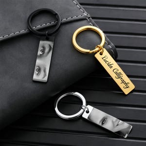 Puede incluir: Tres llaveros con colgantes rectangulares. Un llavero negro con una imagen de ojos. Un llavero dorado con el texto "Lucida Calligraphy". Un llavero plateado con una imagen de una cara sonriente.