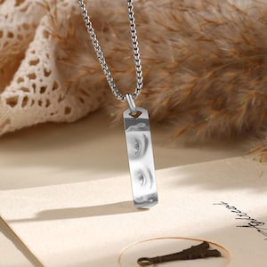 Puede incluir: Collar plateado con un colgante rectangular. El colgante presenta una imagen en blanco y negro de dos ojos. La cadena tiene un diseño pequeño y enlazado. El fondo tiene tonos beige y marrón.