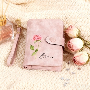 Puede incluir: Cartera rosa con una ilustración de rosa y el nombre "Borrie" impreso. Tiene un cierre de botón a presión y una correa de muñeca. Rosas rosas secas y cristales decorativos en el fondo.