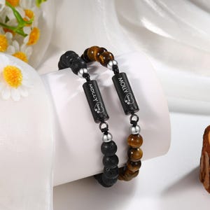 Puede incluir: Dos pulseras a juego. Una presenta cuentas de lava negra, la otra cuentas de ojo de tigre. Ambas tienen colgantes cilíndricos negros con el nombre "MOLLY" y una huella de pata en forma de corazón. Cuentas y conectores plateados completan el diseño.