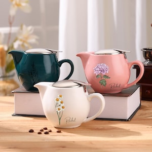 Op de afbeelding: Drie keramische theepotten in teal, roze en wit, elk met een roestvrijstalen deksel en een gebogen handvat. De teal theepot heeft een bloemenontwerp, de roze heeft een paarse bloem en de witte heeft een tros gele bloemen. Elke theepot heeft de naam "Rachel" erop.