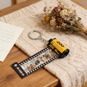 Puede incluir: Un llavero personalizado con diseño de rollo de película. El cilindro amarillo dice "200 36 exp." y está unido a una tira de fotos. El llavero es plateado y descansa sobre una superficie de punto de color crema.