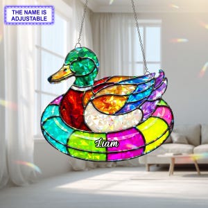 May include: A stained glass duck suncatcher with a green head, red chest, and colorful body. The duck is suspended by a chain and has the name "Liam" on it. The text "THE NAME IS ADJUSTABLE" is in a blue box.