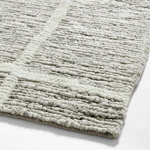 Puede incluir: Primer plano de una alfombra texturizada gris y blanca. La alfombra tiene un pelo grueso y rizado con un diseño geométrico en blanco. Los bordes de la alfombra están acabados con un borde gris. La alfombra está hecha de un material suave y duradero.