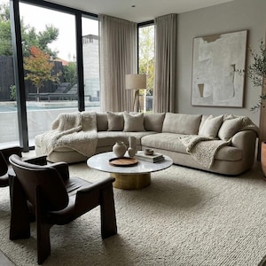 Puede incluir: Salón moderno con un gran sofá seccional beige, dos sillones marrón oscuro y una mesa de centro redonda de mármol. Una gran alfombra cubre el suelo. La habitación tiene grandes ventanales con cortinas beige y una pintura abstracta.