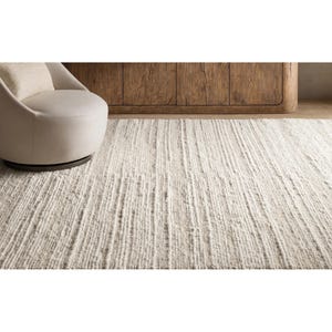 May include: A neutral-toned, textured area rug with a linear pattern. A cream-colored swivel chair with a matching pillow sits on the left. A wooden cabinet is in the background. The rug is a light beige color.