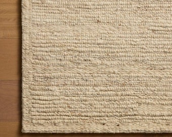 Puede incluir: Una alfombra rectangular con una textura gruesa tejida a mano en un color crema sólido. La superficie muestra filas horizontales de fibras de hilo gruesas y anudadas con variaciones sutiles en el tono y una construcción densa y táctil.
