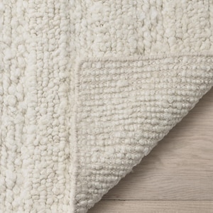 Puede incluir: Alfombra texturizada de color crema con bucles. Una esquina de la alfombra está doblada, revelando la parte inferior con un tejido más apretado. El color neutro y la textura afelpada sugieren un ambiente acogedor.
