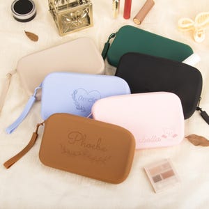 Può includere: Una collezione di trousse rettangolari in vari colori, tra cui beige, blu, marrone, rosa, verde e nero. Alcune trousse presentano nomi e disegni floreali. Ogni trousse ha una chiusura a zip e un cinturino da polso.