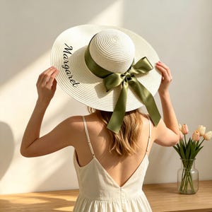 Könnte beinhalten: Cremefarbener Sonnenhut mit breiter Krempe, verziert mit einem grünen Satinband und einer Schleife. Der Name "Margaret" ist in schwarzer Schrift auf dem Rand geschrieben. Ein modisches Accessoire zum Sonnenschutz.