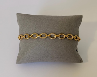 Op de afbeelding: Een goudkleurige kettingarmband bestaande uit een reeks onderling verbonden ovale schakels. Elke individuele schakel vertoont een duidelijke gedraaide of touwachtige textuur op het oppervlak, wat een uniform en herhalend patroon creëert.