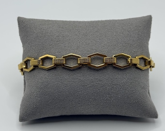 Op de afbeelding: Een goudkleurige armband met een reeks onderling verbonden zeshoekige schakels. Elke schakel wisselt af tussen een gladde, gepolijste afwerking en een getextureerd oppervlak met een kruislingse arcering, wat een geometrisch patroon vormt.