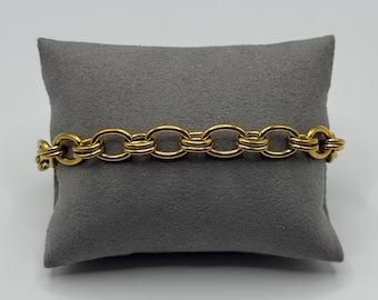 Op de afbeelding: Een goudkleurige kettingarmband met een reeks onderling verbonden ovale schakels. Elke schakel is opgebouwd uit twee parallelle ringen, wat zorgt voor een gelaagd, gestructureerd uiterlijk. De armband ligt horizontaal op een grijs kussentje.