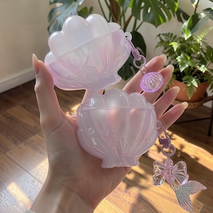 May include: A translucent, iridescent light purple protective case shaped like an open seashell. The case features embossed starfish and bubble details on the surface. It includes a matching purple charm attachment with a small shell and a bow.