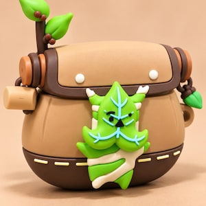 Puede incluir: Una funda protectora de silicona marr&oacute;n para auriculares inal&aacute;mbricos, con forma de peque&ntilde;a mochila con solapa y detalles laterales. Un personaje de color verde brillante con forma de hoja y cara blanca y negra est&aacute; sujeto al frente, y una peque&ntilde;a ramita de hoja verde se encuentra en la parte superior.