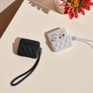 May include: Two quilted earbud cases, one black and one white, each featuring a diamond-stitched pattern and a matching braided wrist strap. The white case is open, revealing a pair of white wireless earbuds inside the charging compartment.
