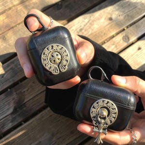 May include: Two black leather protective cases for wireless earbuds, each featuring a circular silver-toned metal emblem with the text CHROME HEARTS and a cross motif. One case includes a metal carabiner, and the other has a dangling cross charm.