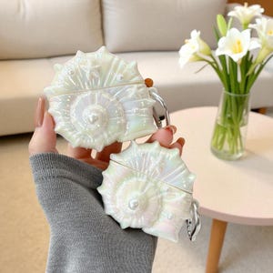 May include: Two iridescent white seashell-shaped cases, possibly for earbuds, are held in a person's hand. Each case has a pearlescent finish, a central pearl, and a silver-coloured clasp. The cases are designed to resemble a seashell.