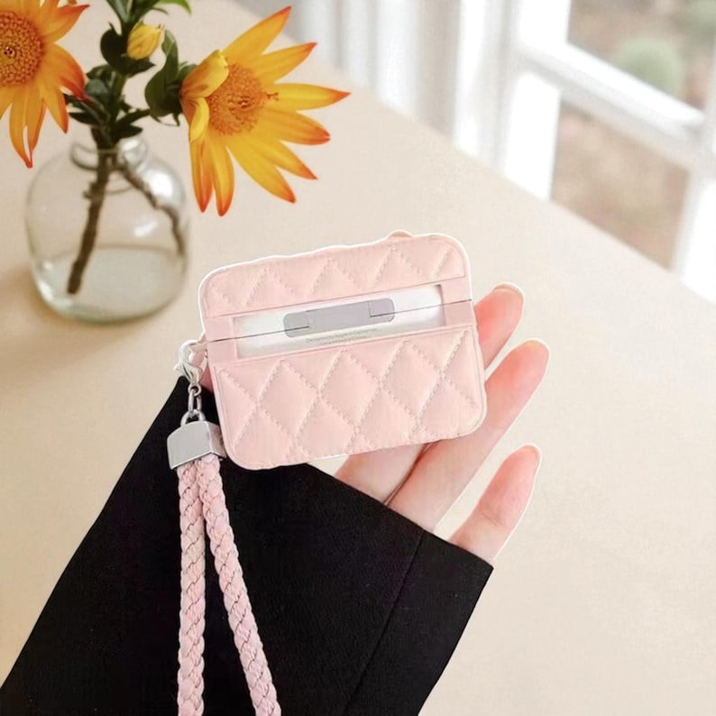 May include: A light pink quilted protective case for wireless earbuds, featuring a diamond-stitched pattern and a matching braided wrist strap attached with a silver-toned metal clasp. The case has a rectangular shape with rounded corners.
