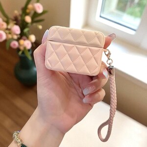 May include: A light pink protective case for wireless earbuds featuring a quilted diamond pattern across the exterior. The case includes a matching braided wrist strap attached to a silver metal clasp on the side, providing a secure way to carry.