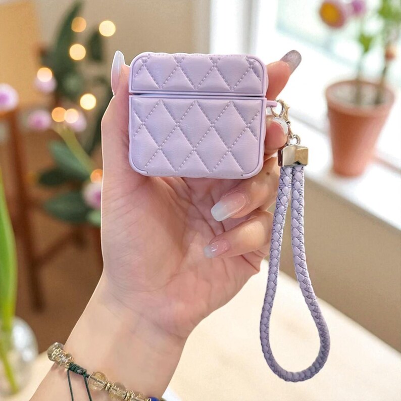 May include: A light purple, quilted protective case for wireless earbuds. The square-shaped case features a diamond-stitched pattern and a matching braided wrist strap attached to a silver-toned metal clasp on the side of the device.