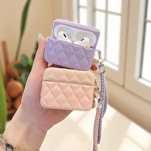 May include: A light purple and a light pink quilted silicone case for wireless earbuds. The purple case is open, revealing white earbuds inside. Both cases feature a diamond-stitched pattern and are attached to a braided purple and pink wrist strap.