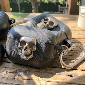 May include: A black carbon fiber textured protective case for wireless earbuds featuring a metallic silver skull emblem on the front and a matching metallic skull on the top lid, equipped with a silver-toned metal carabiner clip attached to the side.